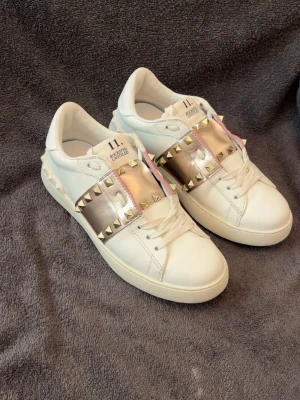 Valentino rockstud 11 - Säljer ett par vita sneakers med breda metallicband i roséguld över vristen, dekorerade med små guldfärgade nitar. Skorna har klassisk låg profil, vita snören och detaljer i rosa längs bandet. Perfekta för dig som vill sticka ut med en edgy och trendig look.