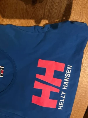 Blå Helly Hansen t-shirt - Snygg blå t-shirt från Helly Hansen med stor röd HH-logga och vit text på bröstet. Klassisk rund halsringning och korta ärmar. Tillverkad i mjuk bomull som känns skön mot huden. Perfekt för en avslappnad och sportig stil.