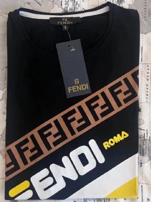 Svart Fendi t-shirt med logga och ränder - Cool svart t-shirt från Fendi med diagonal logga och texten 'Fendi Roma' i vitt och gult. Brunt band med Fendi-monogram går tvärs över bröstet. T-shirten har korta ärmar och är gjord i mjuk bomull. Perfekt för dig som gillar streetstyle och designerplagg.