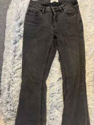 Grå bootcut jeans från Lindex - Snygga grå bootcut jeans från Lindex med klassisk femficksmodell och dragkedjegylf. Jeansen har bälteshällor och är tillverkade i ett mjukt denimtyg som ger en avslappnad look. Perfekta till vardags för en trendig stil.