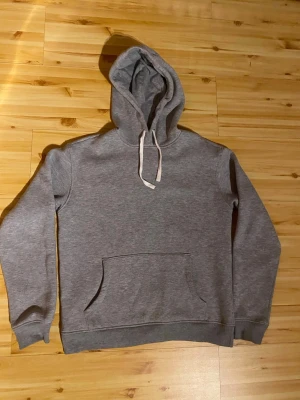 Grå hoodie med biltryck Maggiore - Snygg grå hoodie med huva och vita dragsnören. På ryggen finns ett stort svartvitt tryck av en racingbil och texten 'MAGGIORE'. Klassisk känguruficka framtill och ribbade muddar vid ärmar och midja. Perfekt för dig som gillar streetwear och motorsport.