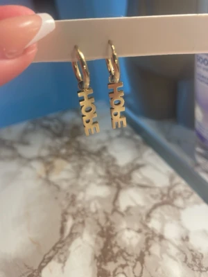 Guldiga HOPE örhängen i metall - Snygga örhängen i guldfärgad metall med texten 'HOPE' som hängsmycke. De har en enkel ringdesign och bokstäverna ger ett coolt statement. Perfekta för dig som vill ha något trendigt och personligt i din stil.