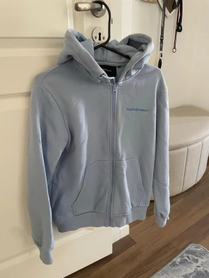 Ljusblå hoodie från Peak Performance - Ljusblå hoodie med dragkedja från Peak Performance. Klassisk modell med huva, två stora fickor framtill och ribbade muddar vid ärmslut och nederkant.  Knappt använd.