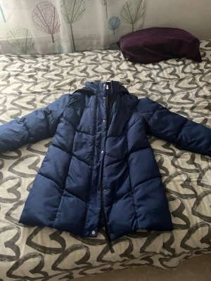 Mörkblå lång pufferjacka med huva - Snygg mörkblå pufferjacka med huva och dragkedja samt tryckknappar framtill. Jackan är lång och har stora quiltade partier för extra värme. Fodret är svart och jackan har en innerficka. Perfekt för kalla vinterdagar.