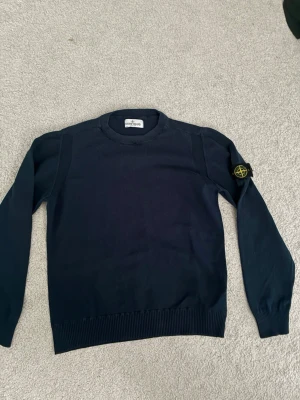 Mörkblå stickad tröja Stone Island - Snygg mörkblå stickad tröja från Stone Island med den klassiska logotypen på vänster ärm. Tröjan har rund halsringning, ribbade muddar och är tillverkad i ett mjukt stickat material. Perfekt för dig som gillar streetwear och vill ha något stilrent. Tröjan är köpt för 2800 kr. Tröjan är lite sönder vid halsen men inget man märker av vid användning. Tröjan passar dig som är runt 152/158. Hör av er vid minsta fråga🥰