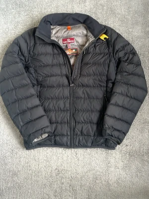 Svart dunjacka från Parajumpers - Snygg svart dunjacka från Parajumpers i modellen Super Lightweight. Jackan har quiltad design, hög krage och dragkedja framtill. Insidan är ljusgrå och jackan har en gul detalj vid halsen. Perfekt för kalla vinterdagar. Passar dig mellan 165 till 175