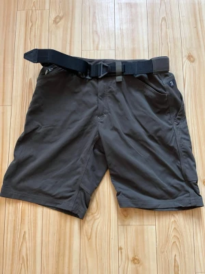 Bruna shorts från Haglöfs med bälte - Säljer ett par bruna shorts från Haglöfs i storlek XL. De har ett praktiskt svart bälte i midjan och dragkedja vid fickorna. Perfekta för utomhusaktiviteter och har en loose passform. Materialet känns slitstarkt och bekvämt.