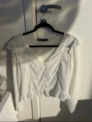 Vit blus med volanger  - Söt vit blus från Zara med prickigt mönster, djup V-ringning och dekorativa volanger vid axlar och nederkant. Blusen har spetsdetaljer längs framsidan och långa ärmar i ett lätt, transparent tyg. Perfekt för en trendig och romantisk look.