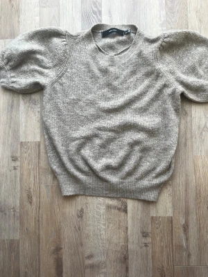 Beige stickad tröja med puffärm - Snygg beige stickad tröja med puffiga korta ärmar och rund hals. Tröjan har ribbade muddar vid ärmslut och nederkant. Perfekt för dig som vill ha en trendig och mysig look till hösten.