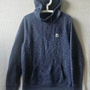 Mörkblå hoodie från Hollister - Snygg mörkblå hoodie från Hollister med melerad look och klassisk logga på bröstet. Hoodien har huva med dragsko och en stor magficka framtill. Perfekt för en avslappnad och trendig stil.
