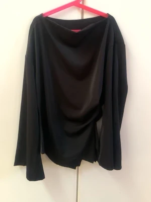 Svart långärmad topp med vid hals - Säljer en svart långärmad topp med lös passform och vid, draperad halsringning. Tröjan har vida ärmar och är gjord i ett mjukt, tunt material som faller snyggt. Perfekt för dig som gillar en avslappnad men ändå stilren look.