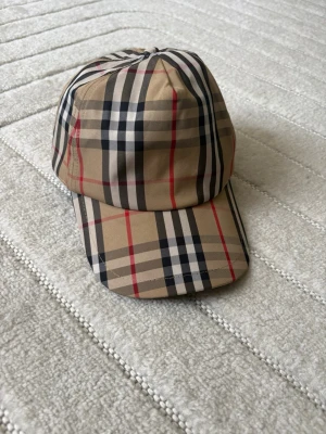 Beige keps med rutigt mönster - Snygg beige keps med klassiskt rutigt mönster i svart, vitt och rött. Kepsen har böjd skärm och justerbar passform. Perfekt accessoar för att lyfta din outfit med en trendig touch.
