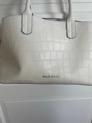 Vit handväska i crocopräglat skinn - Snygg vit handväska från Valentino bags (Mario Valentino) med crocopräglat mönster och två rejäla handtag. Väskan har en stilren och rymlig design, perfekt för dig som gillar klassiska accessoarer med en twist. Materialet är konstläder med struktur och väskan har guldfärgad logga framtill. Får plats med ex skoldator.