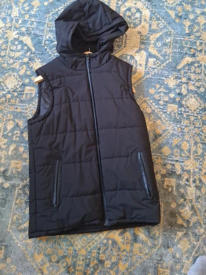 Svart dunväst med huva - Snygg svart dunväst med huva och dragkedja framtill. Västen har två praktiska fickor med dragkedja och quiltad design. Perfekt för lager på lager och ger en clean look till din outfit.