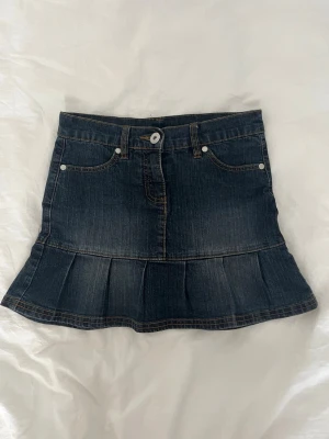 Mörkblå jeanskjol med volang - Säljer en mörkblå jeanskjol i kort modell med volang nedtill. Kjolen har klassiska fickor fram, bälteshällor och stängs med knapp och dragkedja. Perfekt för dig som gillar denim och vill ha en snygg och enkel kjol till sommaren.