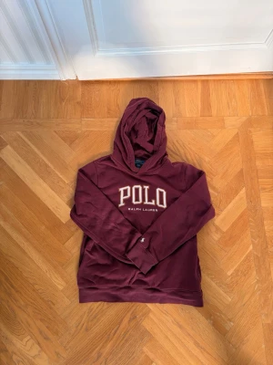 Vinröd barn hoodie från Polo Ralph Lauren - Snygg vinröd hoodie från Polo Ralph Lauren med stor POLO-logga broderad på bröstet. Klassisk huva och mjukt material som känns skönt mot huden. Perfekt för dig som vill ha en stilren och bekväm hoodie med streetwear-vibe.