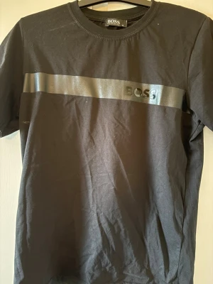 Hugo boss T-shirt  - Snygg svart t-shirt från BOSS med ett stilrent blankt band och BOSS-logga över bröstet. Klassisk rund halsringning och korta ärmar. Tillverkad i mjuk bomull för skön känsla och enkel passform.