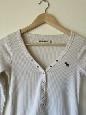 Vit ribbad långärmad topp Abercrombie & Fitch - Vit ribbad långärmad topp från Abercrombie & Fitch med v-ringning och knappar framtill. Liten blå logga broderad på bröstet. Stretchigt material och figurnära passform, perfekt till jeans eller kjol.