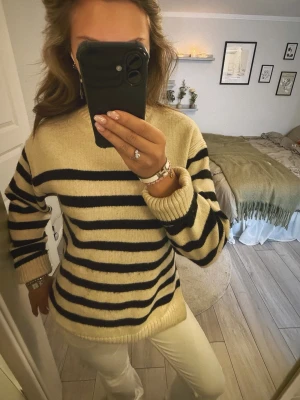 Beige stickad tröja med svarta ränder - Säljer en beige stickad tröja med svarta ränder. Tröjan har en rak passform, halterneck och ribbade muddar vid ärmslut och nederkant. Perfekt för dig som gillar klassisk och avslappnad stil.