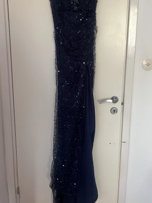 Mörkblå glittrig långklänning - Säljer en elegant mörkblå balklänning med glittriga paljettdetaljer. Klänningen har en åtsittande passform och ärmarna är axelbandslösa. Perfekt för bal eller festliga tillfällen.
