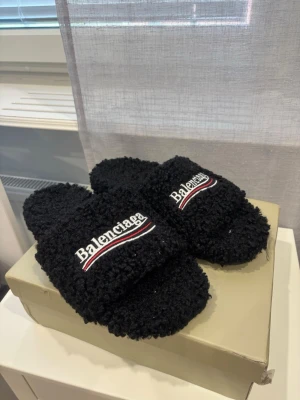 Svarta fluffiga tofflor  - Säljer ett par svarta, fluffiga tofflor med öppen tå och bred rem. Tofflorna har broderad logga i vitt, rött och svart på ovansidan. Perfekta för dig som vill ha något riktigt cozy och trendigt hemma.