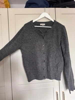 Mörkgrå stickad kofta - Mysig mörkgrå stickad kofta med rund halsringning och knappar framtill. Koftan har långa ärmar och ribbade muddar vid ärmslut och nederkant. Perfekt att slänga över en t-shirt för en avslappnad look. Storlek L men sitter som en S