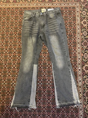 Gråa jeans från Gallery Dept. - Unika gråa jeans från Gallery Dept. med slitna detaljer och ljusare paneler längs benen. Modellen har raka ben med lätt utsvängning och råa kanter nertill. Klassisk femficksdesign och knappgylf. Perfekta för dig som gillar streetwear och statement-plagg.