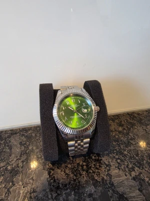 Ditt pris! - Stilren klocka i silverfärg med metallarmband och en grön urtavla. Klockan har tydliga siffror, datumvisning och en räfflad kant runt urtavlan. Perfekt accessoar för dig som vill sticka ut med en unik färg på handleden.