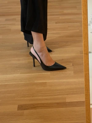 Svarta slingback pumps med spetsig tå - Säljer ett par svarta pumps med smal klack och  slingback. Skorna har spetsig tå och en elegant, stilren design. Nyskick, endast använt inhomhus för plåtning.