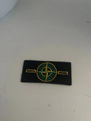 Patch stone Island  - Säljer ett klassiskt Stone Island tygmärke i svart med broderad gul och grön kompasslogga. Märket är rektangulärt och har detaljer i gult och grönt, perfekt att fästa på jacka eller tröja för en streetwear-look.