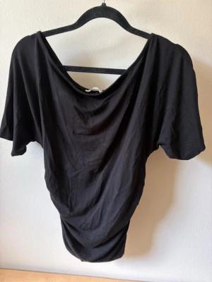 Svart loose topp med vid hals - En svart topp med lös passform och tajt runt magen , draperad halsringning. Toppen har korta ärmar och är tillverkad i ett mjukt tyg som faller snyggt. Perfekt för en avslappnad och trendig look.