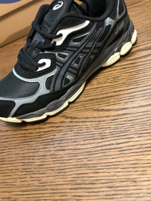 Svarta och grå sneakers acics gel - Snygga svarta och grå sneakers från Asics med klassisk logga på sidan och GEL-dämpning i sulan. Ovandelen är i mesh och syntetmaterial för bra andningsförmåga och komfort. Sulan är vit och svart med tydlig struktur. Perfekta för en sportig och trendig look.