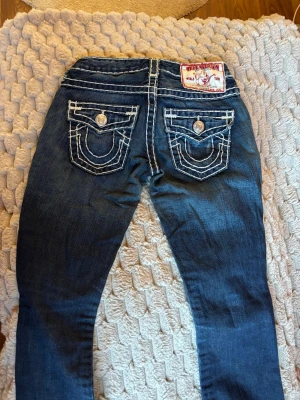 Mörkblå True Religion jeans - Säljer ett par mörkblå jeans från True Religion med vita kontrastsömmar och ikoniska fickdetaljer bak. Storlek W23. Klassisk femficksmodell med raka ben och snygga knappar. Perfekta för dig som vill ha en streetig och trendig look.