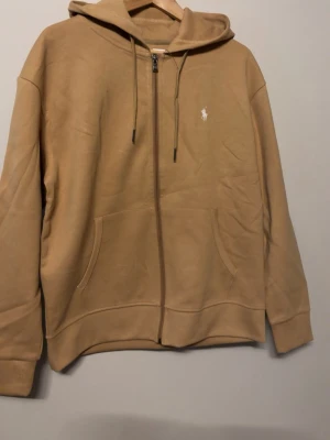 Beige hoodie med dragkedja och huva - Säljer en beige hoodie med dragkedja, huva och snörning. Den har fickor framtill och en liten vit logga på bröstet. Hoodien är långärmad och har en relaxed passform, perfekt för chill dagar.