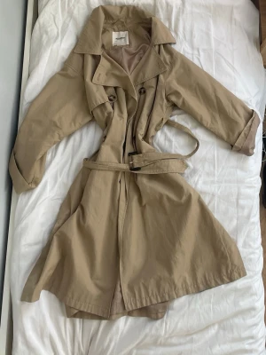 Beige trenchcoat från Pull&Bear - Stilren beige trenchcoat från Pull&Bear med klassisk dubbelknäppning, bred krage och avtagbart midjebälte. Lätt material som ger en snygg siluett och passar perfekt till lager-på-lager. Fodrad insida och långa ärmar för extra komfort. Obs: belt clasp is broken
