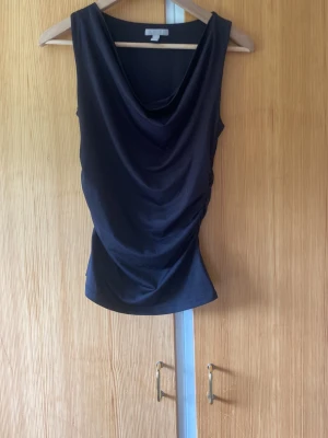 Svart ärmlös topp med drapering - Snygg svart ärmlös topp med draperad framsida och djupare ringning. Toppen har en smickrande passform och är tillverkad i ett mjukt, stretchigt material som känns bekvämt mot huden. Perfekt att styla med jeans eller kjol för en trendig look.