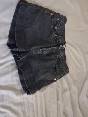 Svarta denimshorts med hög midja - Säljer ett par svarta denimshorts med hög midja och uppvikta benslut. Shortsen har klassisk femficksdesign, kopparfärgade nitar och knappar framtill. Perfekta för sommaren och ger en avslappnad vibe till din outfit.