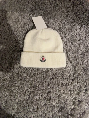 Vit ribbstickad mössa Moncler - Snygg vit ribbstickad mössa från Moncler med uppvikt kant och klassisk logotyp framtill. Tillverkad i mjukt material som håller dig varm och stilren under kalla dagar. Enkel och clean design som passar till det mesta.