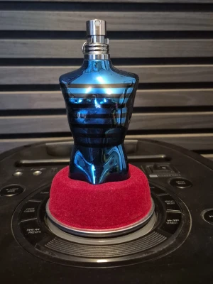 Blå parfymflaska i form av torso - Unik parfymflaska i metalliskt blått glas formad som en manlig torso med svarta ränder. Flaskan har en sprayfunktion i silverfärg och står på en röd sammetsliknande bas. Perfekt som snygg inredningsdetalj eller till din doftsamling. Den är halv full. Så ca 30-40 ml kvar