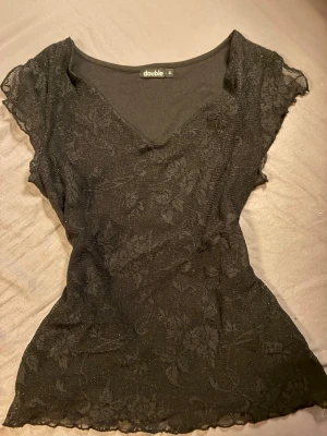 Svart glittrig topp från Double - Snygg svart topp från Double med glittrigt tyg och diskret blommönster. Toppen har v-ringning och korta ärmar, perfekt för dig som vill ha något extra till outfiten. Materialet känns lätt och stretchigt. OBS att midjan är lite intryckt för bilden. Storlek medium.  