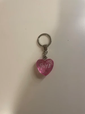 Nyckelring hjärta med glitter - Säljer en söt nyckelring med ett rosa, glittrigt hjärta i plast. På hjärtat står namnet 'Elvira' skrivet i vitt. Perfekt att hänga på väskan eller nyckelknippan för en personlig touch.