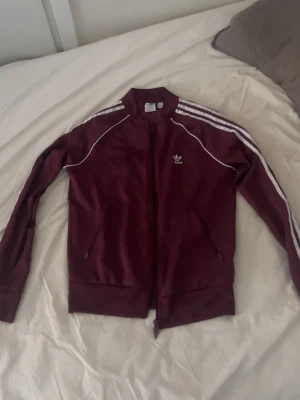 Vinröd Adidas track jacket - Säljer en vinröd Adidas track jacket med vita ränder på ärmarna och dragkedja framtill. Jackan har två fickor med dragkedja och är tillverkad i Primeblue-material som innehåller återvunnen plast från havet. Klassisk Adidas-logga på bröstet.