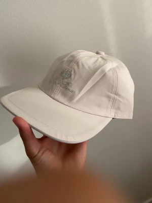 Beige keps från Loro Piana - Snygg beige keps från Loro Piana med broderad logga framtill. Kepsen har böjd skärm och är tillverkad i ett slitstarkt material med Storm System-teknologi. Insidan har rutigt foder och röd kant. Perfekt för dig som vill ha en stilren och exklusiv look.