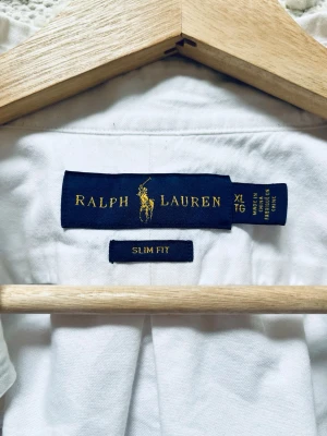 Vit slim fit skjorta från Ralph Lauren - Klassisk vit skjorta från Ralph Lauren i slim fit-modell. Skjortan har lång ärm, knappar framtill och den ikoniska broderade loggan på bröstet. Tillverkad i mjuk bomull och passar perfekt till både jeans och chinos.