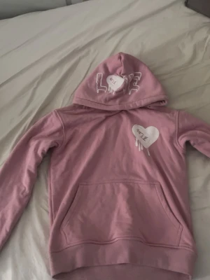 Rosa hoodie med hjärtdetalj - Säljer en rosa hoodie med huva och magficka. På bröstet och huvan finns vita tryck med texten 'LOVE' och ett hjärta som ser ut att smälta. Hoodien har ribbade muddar och en avslappnad passform. Perfekt för en chill och trendig look.
