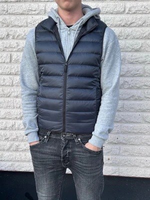 Blå dunväst med dragkedja - Snygg svart dunväst med quiltad design och dragkedja framtill. Västen har en klassisk, rak passform och är perfekt att bära över en hoodie. Tillverkad i ett glansigt material som ger en modern look.