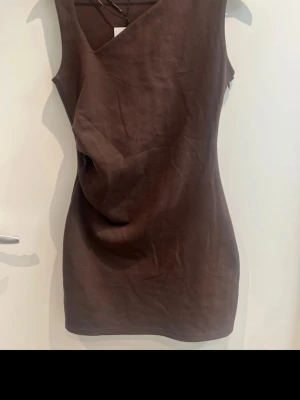 Brun ärmlös klänning - Säljer en brun ärmlös klänning med snygg drapering framtill och v-ringad hals. Klänningen har en kroppsnära passform och är tillverkad i ett mjukt material som känns bekvämt mot huden. Perfekt för dig som gillar stilrena och moderna plagg.