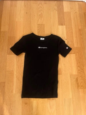 Svart Champion t-shirt - Svart t-shirt från Champion med vit logga på bröstet och liten logga på ärmen. Klassisk passform och rund halsringning. Perfekt för en avslappnad och sportig stil.