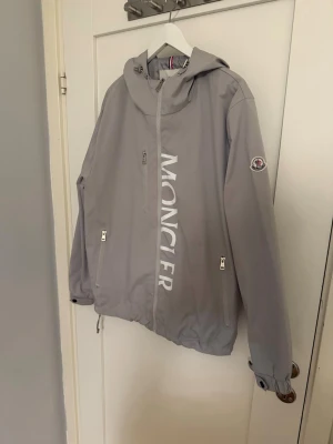 Grå vindjacka med huva från Moncler - Snygg Windbreaker från Moncler i storleken XL men de är mindre i storleken. Därför passar den bättre för storlek L. 