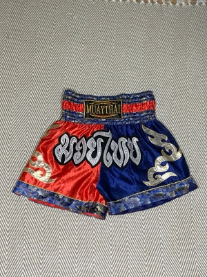Muay Thai shorts röd/blå med mönster - Säljer ett par Muay Thai shorts i blankt material med ena sidan röd och andra sidan blå. Shortsen har gulddetaljer, thailändska tecken och bred resår i midjan. Dekorativa mönster i guld och blått längs kanterna. Perfekta för kampsportsträning.